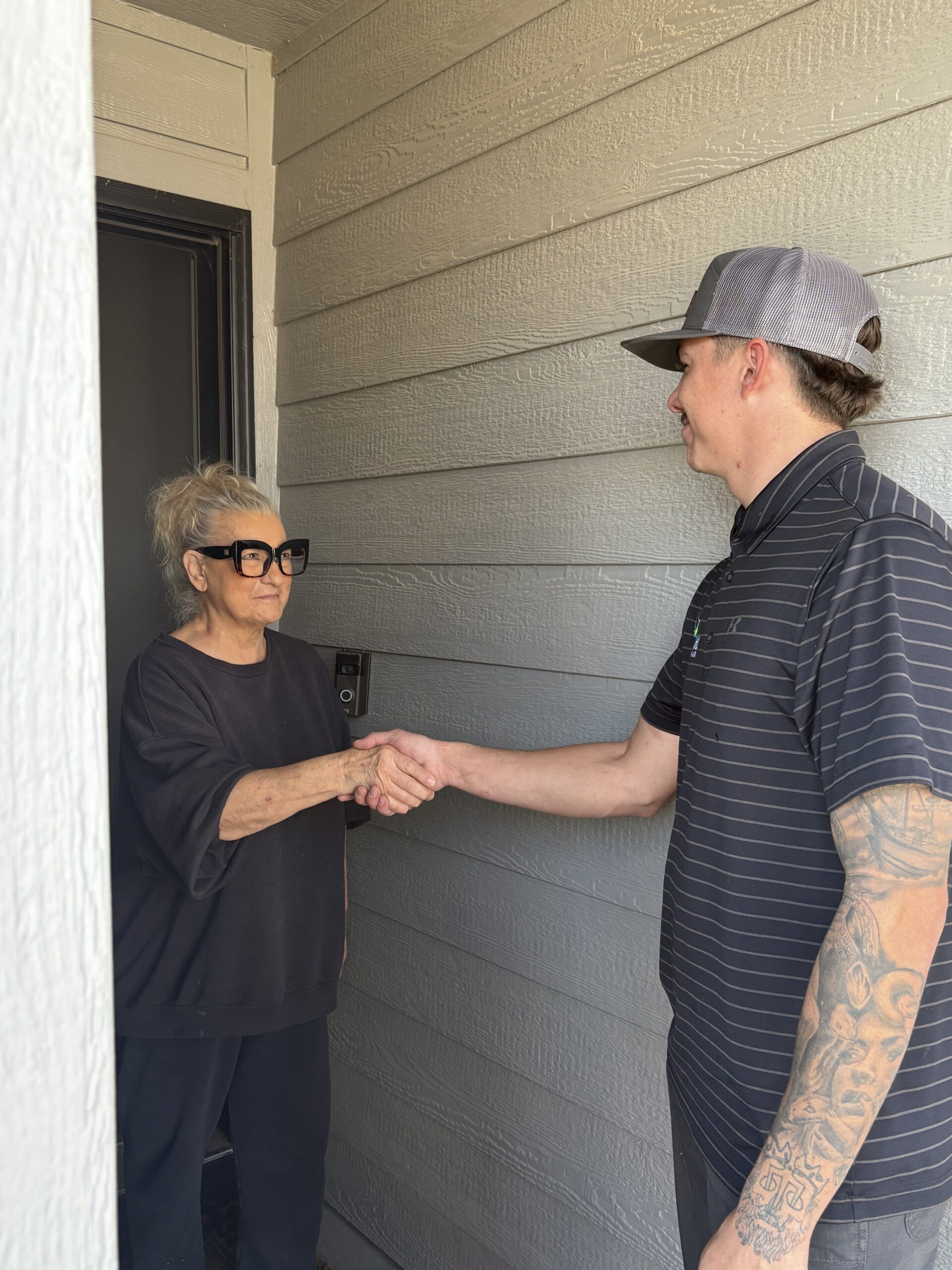 A woman and a man shaking hands at the doorway of a house. The woman stands inside, while the man stands outside, wearing a cap and short-sleeved shirt.