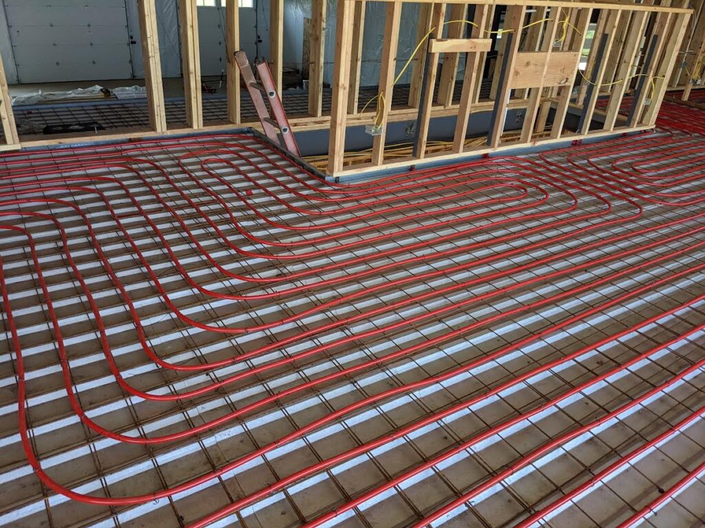 Red radiant floor heating pipes laid out on a grid across the subfloor of a house under construction, with exposed wooden framing and electrical wiring visible.