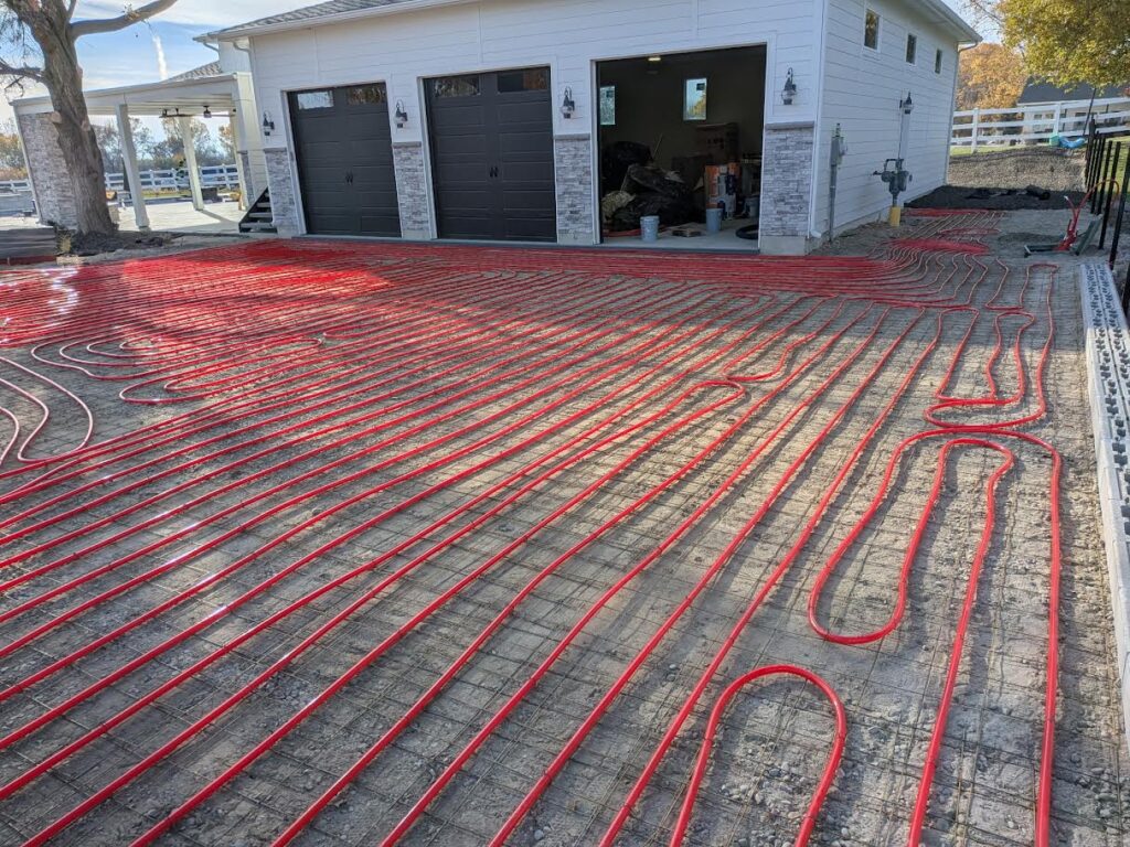 Red radiant heating tubes are installed in a grid pattern on the ground in front of a white garage with black doors, preparing for a heated driveway system.