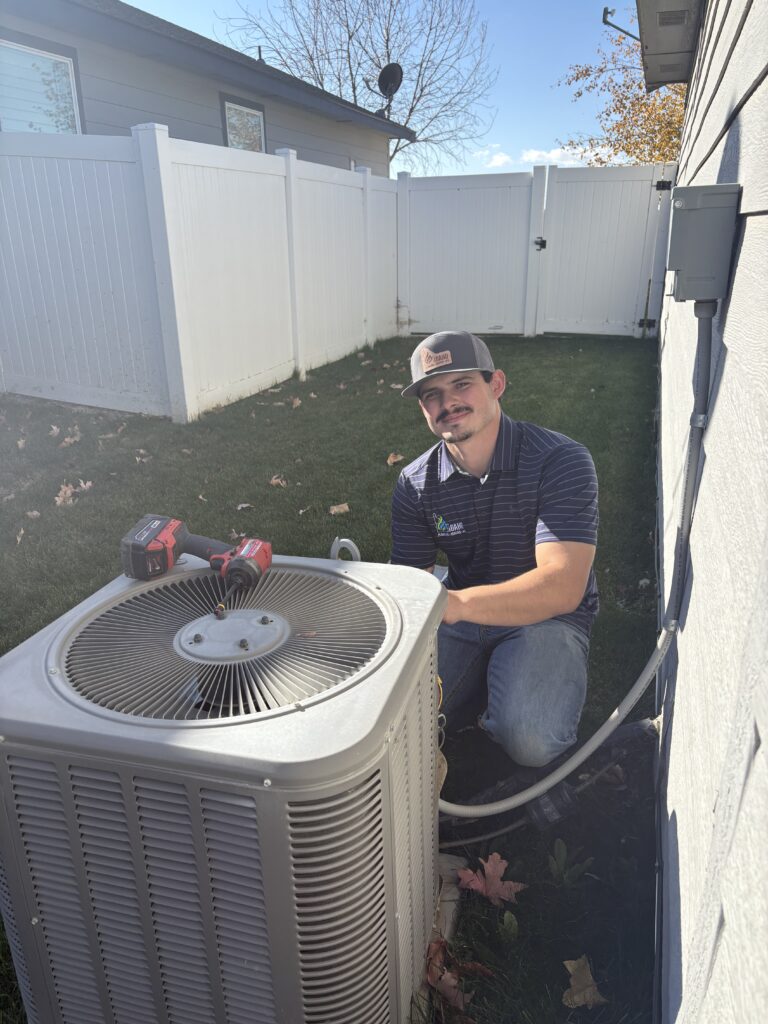 A technician kneels next to an outdoor air conditioning unit, holding a tool, with more equipment resting on top of the unit.
