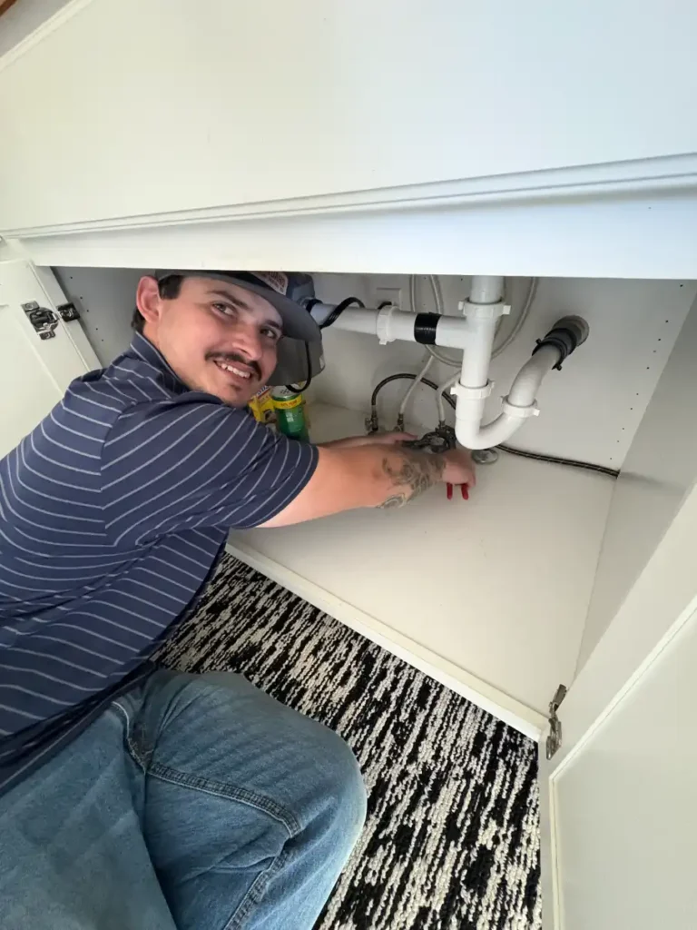 A person in a striped shirt is crouched under a sink, working on the plumbing with tools.